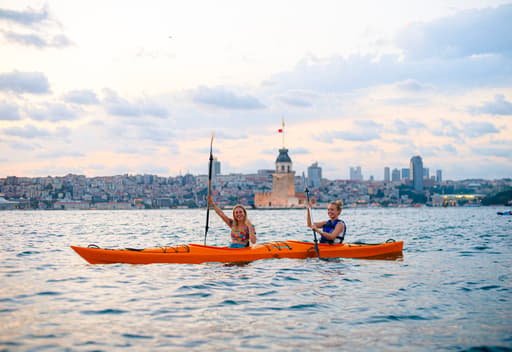 Gülümseyen iki kadın turuncu ikili kanoda küreklerini kaldırıyor, İstanbul Boğazı'nda Kız Kulesi önünde kano