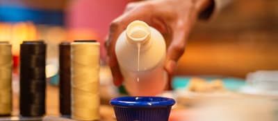 Hand pouring adhesive into a blue dish beside thread spools at a leather craft workshop in Istanbul.