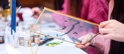 Hands holding a brass-framed glass panel with black Arabic calligraphy at a calligraphy workshop in Istanbul.