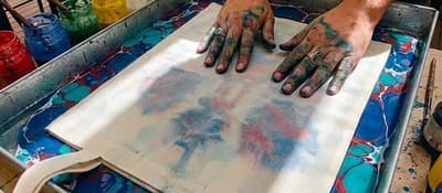 Hands press paper onto a red-and-blue marbling tray at an Ebru workshop in Istanbul.